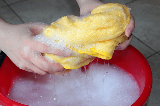 Hands washing a cleaning cloth in a bucket of soapy water, demonstrating gentle spring cleaning for outdoor pool furniture maintenance.